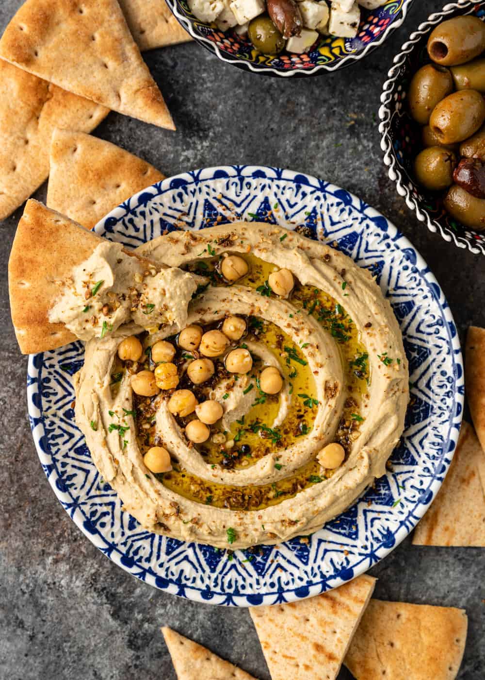 close up image of garbanzo bean hummus on a pita chip lying on the edge of bowl full of the dip