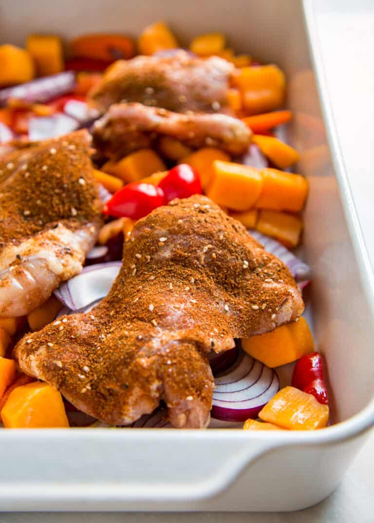 african spices on raw chicken pieces in white baking dish with carrots, red onion, peppers