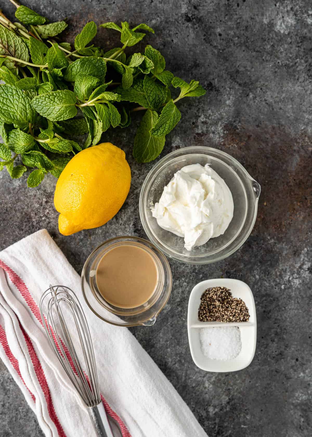 overhead image: bowl of Greek yogurt, whole fresh lemon, fresh mint, tahini paste, and salt and pepper, all on a gray backdrop with a whisk and kitchen towel