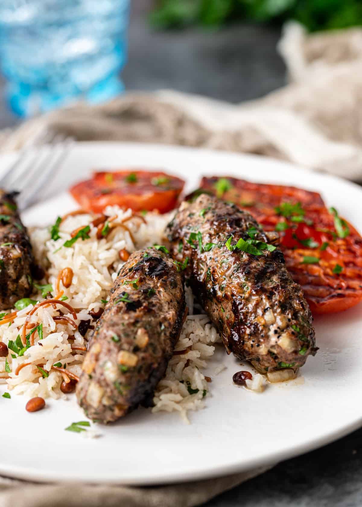 grilled ground meat formed into log shapes served on white plate with rice and grilled tomatoes