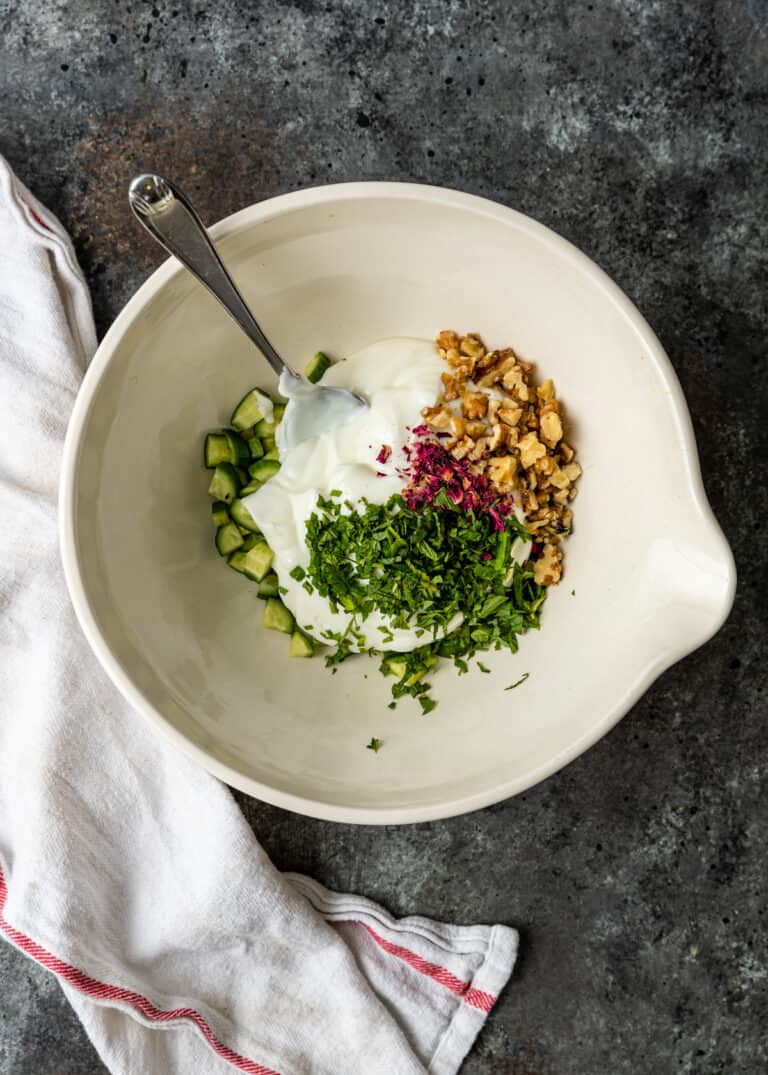 overhead image: diced Persian cucumbers, walnuts, rose petals and yoghurt in white bowl for salad