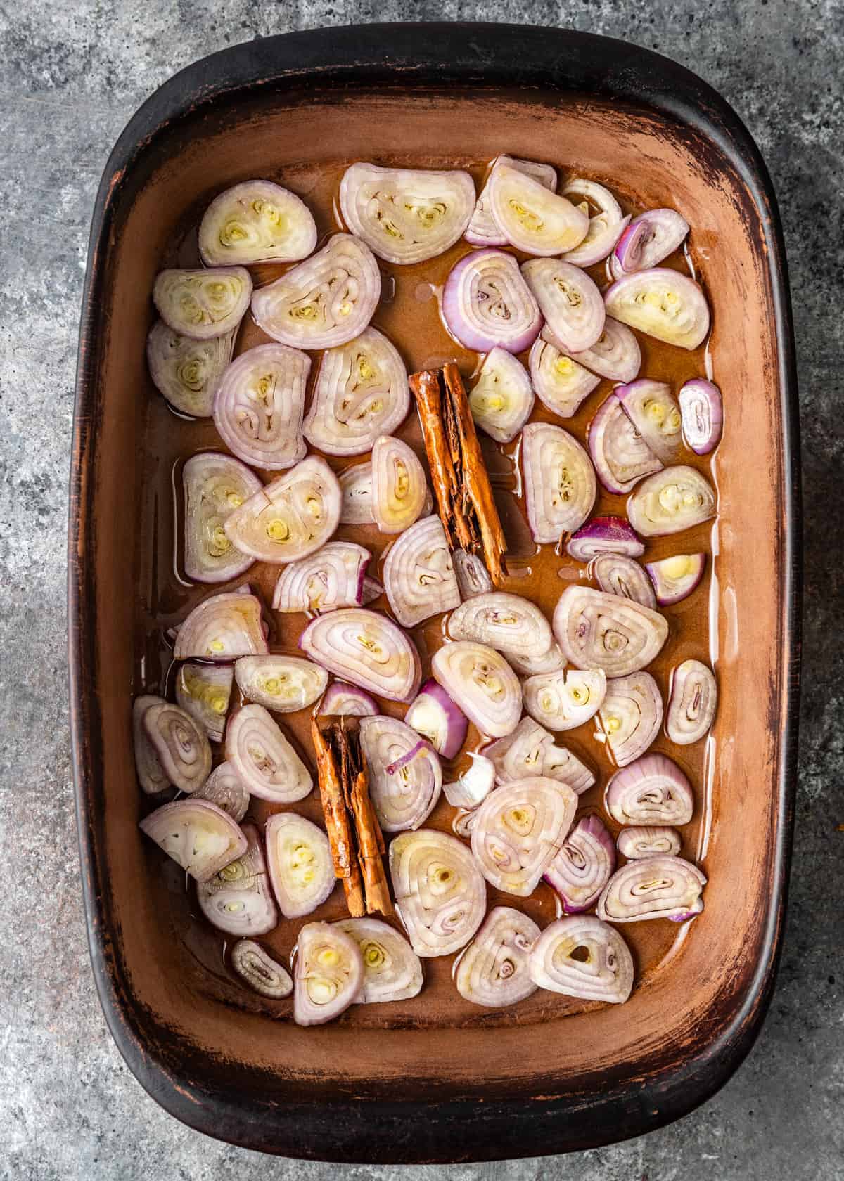 overhead: thin slices of shallot in baking dish with cinnamon sticks, ready for oven roasting