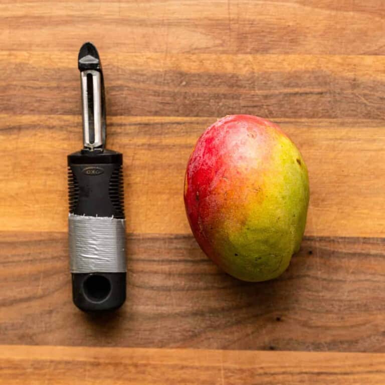 whole fresh mango on cutting board with vegetable peeler