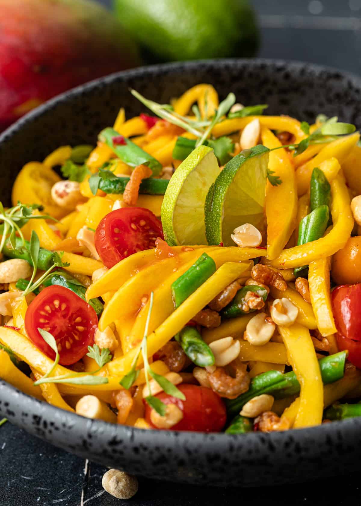 side view close up: green mango salad with mango fruit, tomatoes, beans, and peanuts in black bowl