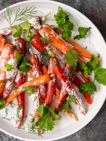 overhead: round white plate of Moroccan carrots with yogurt sauce and garnish of fresh dill and cilantro