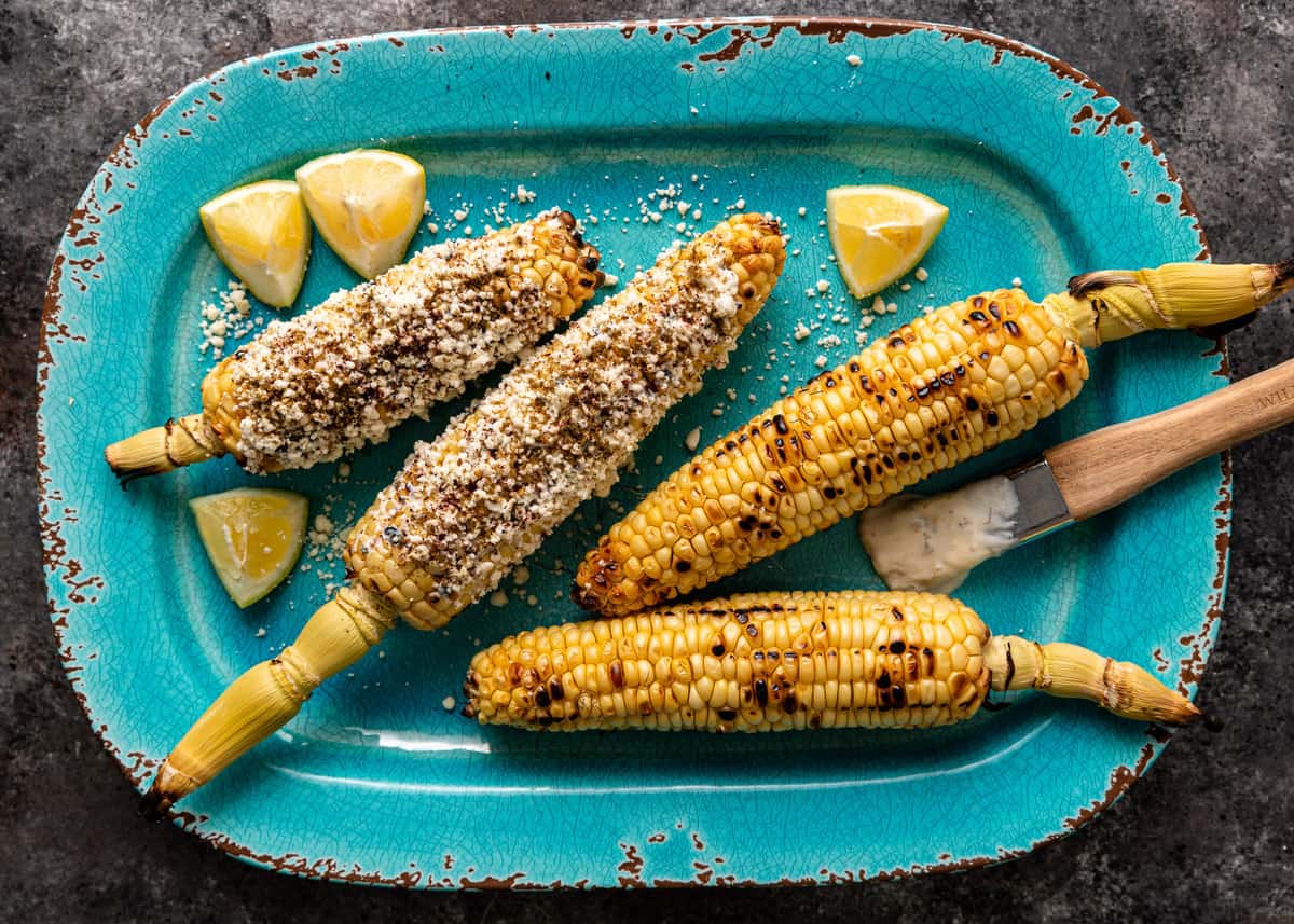overhead: a blue platter with street corn and lemon wedges and a brush with yogurt sauce