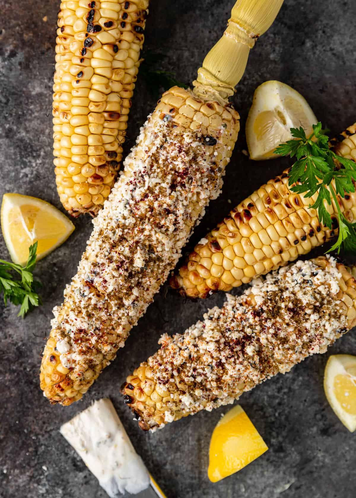 overhead: Mediterranean street corn with feta and seasonings on a countertop with lemon wedges and fresh herbs