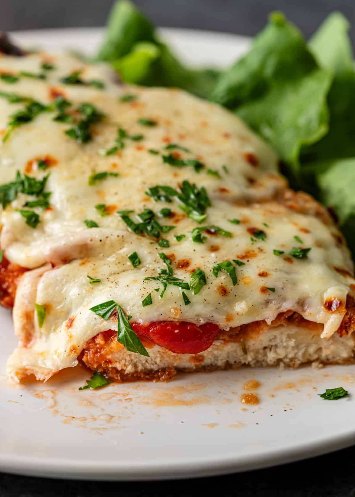 closeup: chicken parmigiana cut on a plate so that the chicken is visible. It's covered with marinara, cheese, and fresh herbs