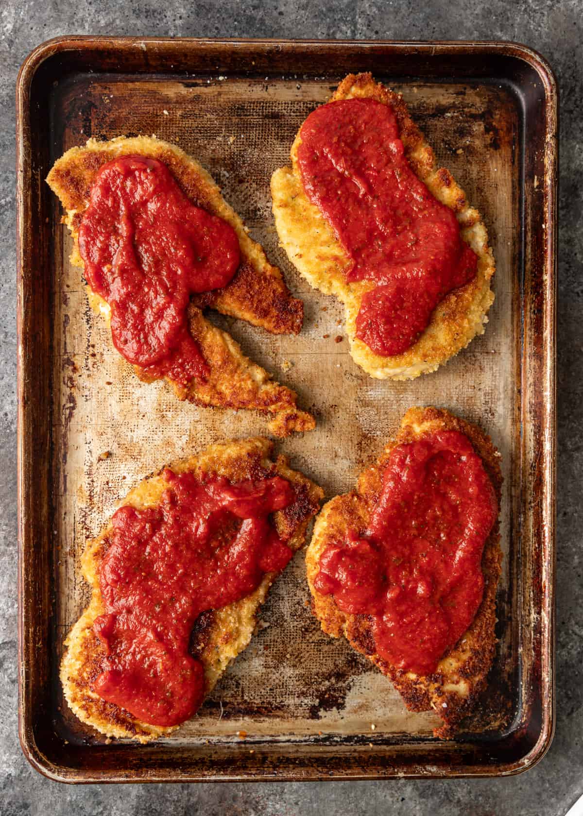 overhead: cooked breaded chicken breasts on a baking sheet with marinara on top