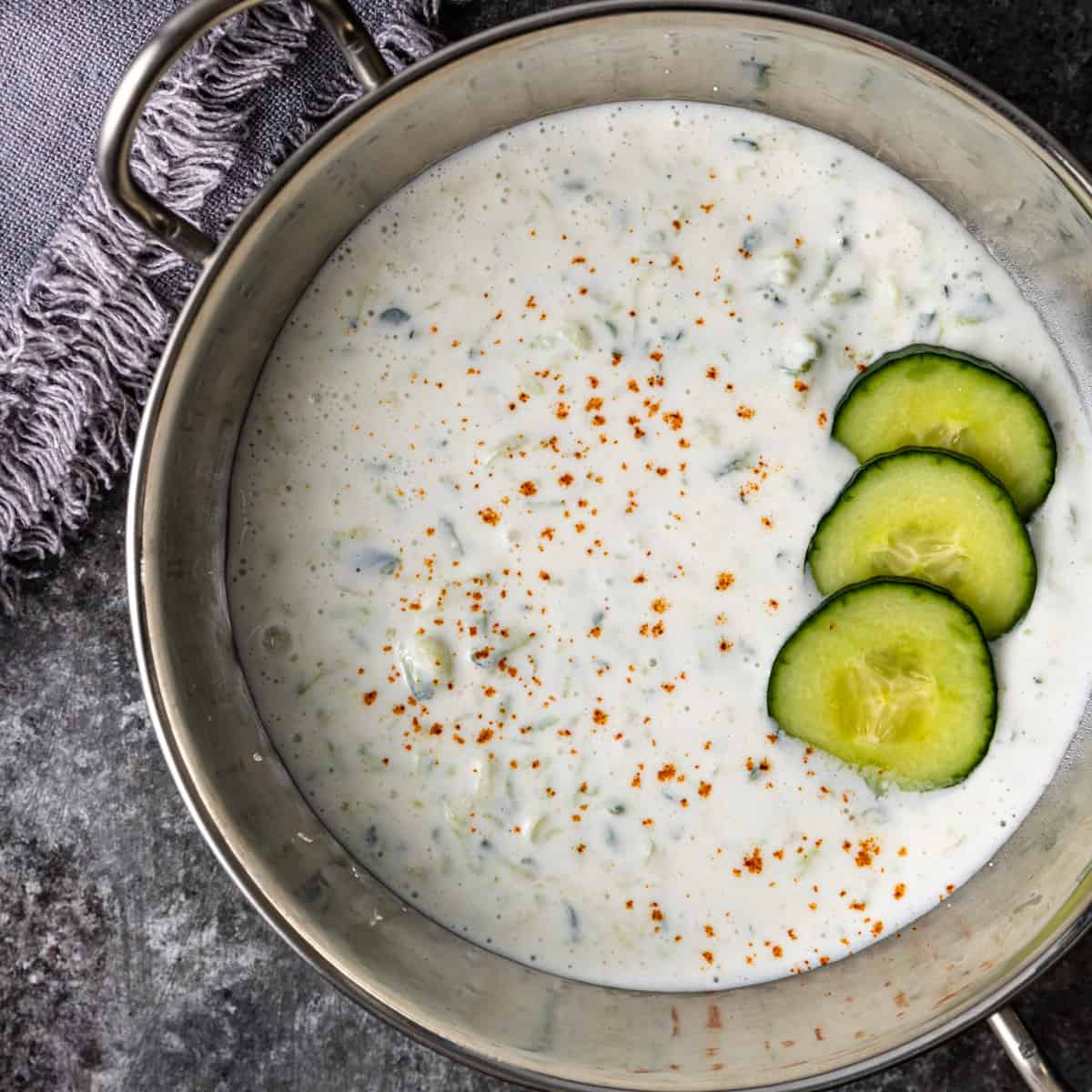 overhead: cucumber raita in a silver bowl with sliced cucumbers on top