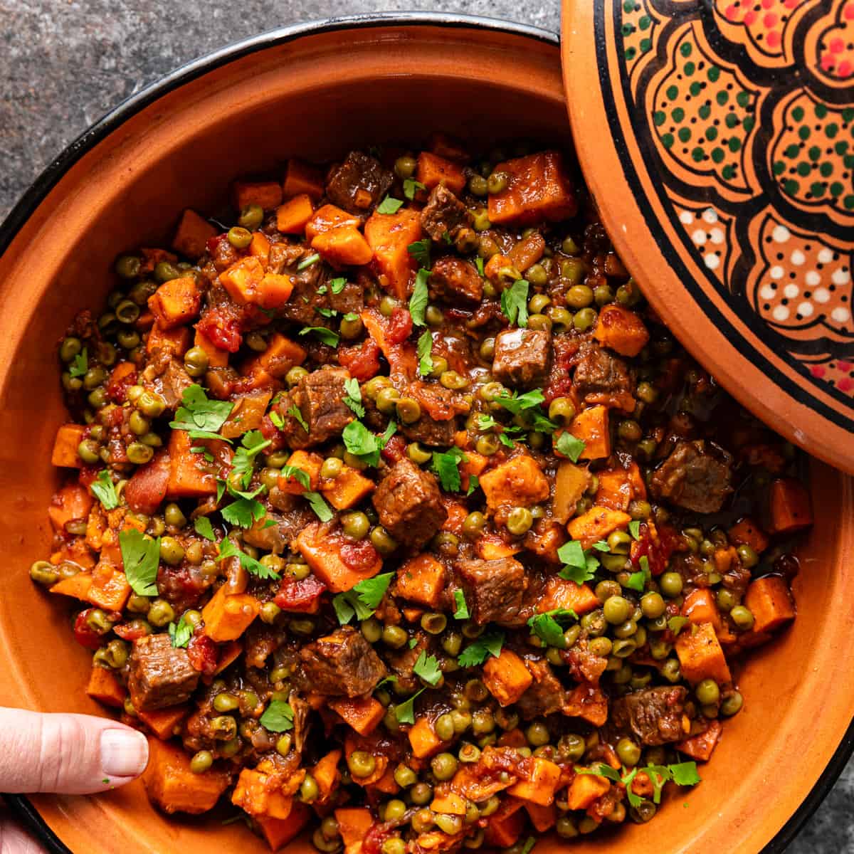overhead: a hand holding a large dish of beef tagine with sweet potatoes and peas